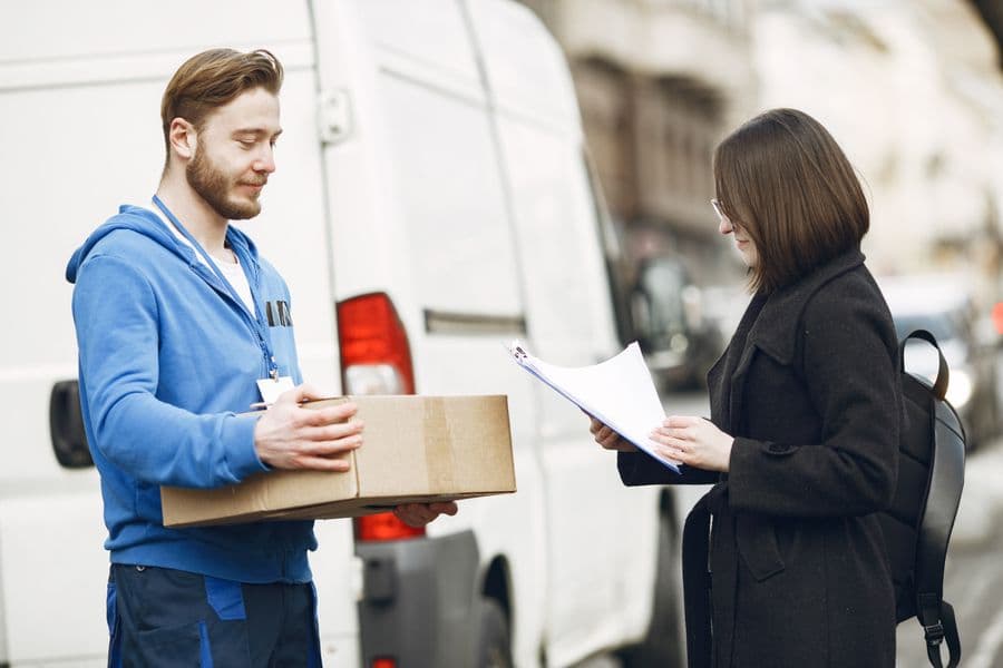 A courier handing a parcel to a customer beside a delivery van while she signs for it.